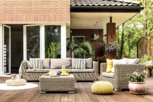 plan early for outdoor living spaces in 2022 Yellow pouf next to a rattan armchair and flowers on wooden patio with striped pillows on sofa