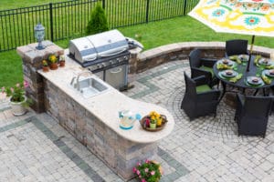 outdoor kitchens - High angle view of a stylish outdoor kitchen, gas barbecue and dining table set for entertaining guests with formal place settings and flowers on a paved patio